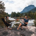 2 personer ved elva som setter på hver sin camp stol med bord i mellom norse camp