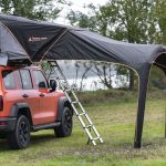 Et oransje terrengkjøretøy med et oppslått taktelt og en montert baldakin som gir skygge på campingplassen.